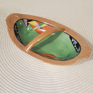 May include: An oval ceramic serving dish with a peach-colored rim and a central arched handle. The interior features a green base with geometric accents in black, yellow, orange, and purple, including triangular patterns along the curved edges.