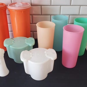 Peut inclure: Ensemble de vaisselle de cuisine en plastique vintage de différentes couleurs. Comprend des gobelets orange, des tasses aux couleurs pastel et des salières et poivrières blanches. Les articles sont disposés sur une surface noire.