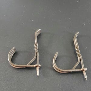 Könnte beinhalten: Zwei rustikale Metall-Wandhaken. Jeder Haken hat ein gebogenes Doppel-Draht-Design mit einem gedrehten Stiel und einer Schraube zur Befestigung. Die Haken haben eine dunkle, gealterte Metallfarbe.
