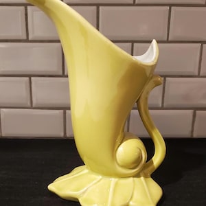 May include: A yellow ceramic vase with a unique, curved design. The vase features a flared top, a decorative handle, and a base shaped like a stylized leaf. The interior is white. A decorative piece for the home.