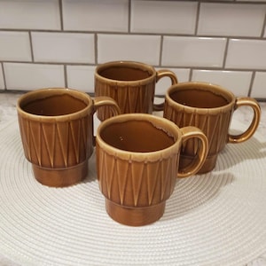 Könnte beinhalten: Vier braune Keramikbecher mit geometrischem Design. Jeder Becher hat einen Henkel und einen leicht ausgestellten Rand. Die Becher sind auf einer weißen, strukturierten Oberfläche mit weißem Fliesenhintergrund angeordnet.