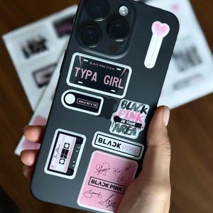 May include: A black phone case adorned with multiple stickers. The stickers feature text like "TYPA GIRL", "BLACK PINK", and a cassette tape design. The case is held in a hand, with other printed materials in the background.