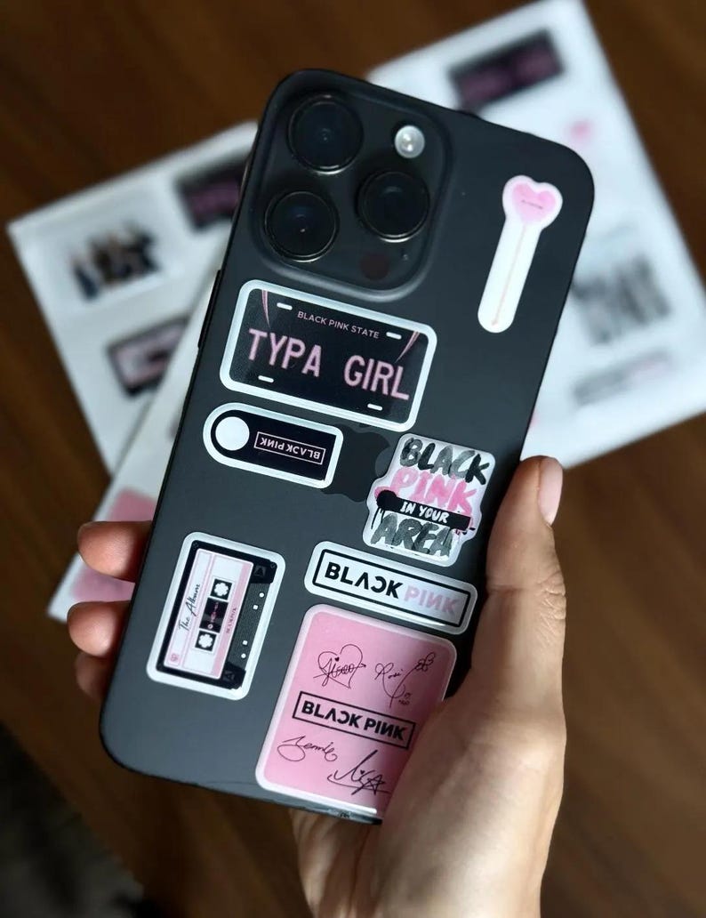 May include: A black phone case adorned with multiple stickers. The stickers feature text like "TYPA GIRL", "BLACK PINK", and a cassette tape design. The case is held in a hand, with other printed materials in the background.
