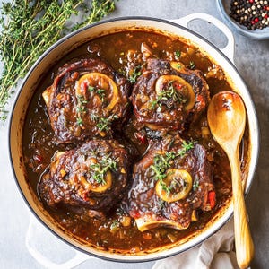 May include: A white ceramic pot filled with a rich, brown stew containing four large pieces of braised beef. The stew is garnished with fresh thyme sprigs and lemon slices. A wooden spoon rests on the edge of the pot.