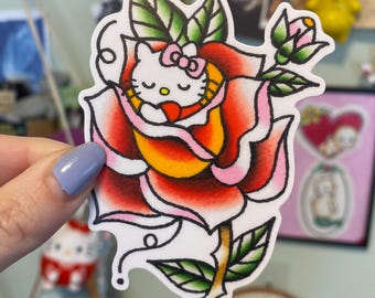 May include: A sticker featuring a Hello Kitty design within a traditional rose tattoo. The rose has red, pink, and yellow petals with green leaves. The sticker is held by a hand with light blue nail polish.