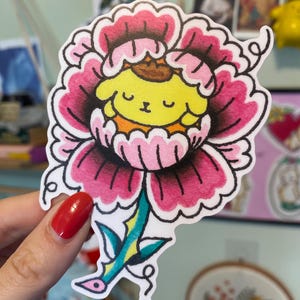 May include: A colorful sticker featuring a cartoon character inside a pink and black flower. The character has yellow fur, a brown hat, and a smiling face. The flower has a green and blue stem.