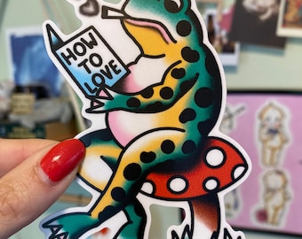 May include: A colorful sticker featuring a frog reading a book titled "HOW TO LOVE." The frog is sitting on a mushroom and smoking, with heart-shaped smoke. The frog has green, yellow, and black markings.