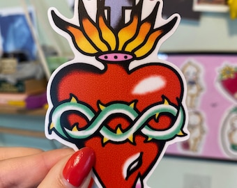 May include: A sticker featuring a red heart with a crown of thorns, a cross, and flames. The heart has a teardrop design and is outlined in black. The sticker is held by a person with red nail polish.
