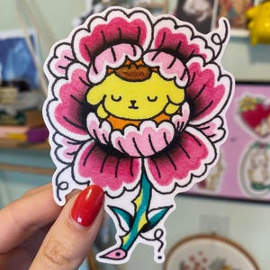 May include: A sticker featuring a yellow cartoon character inside a pink and red flower. The flower has a green and yellow stem with blue accents. The sticker is held by a hand with red nail polish.