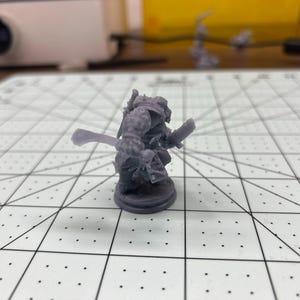 May include: A gray plastic miniature figurine of a warrior holding a sword and shield. The figurine is standing on a round base.