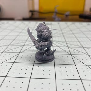 May include: A gray plastic miniature figurine of a fantasy character holding a sword. The figurine is standing on a round base.