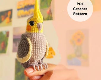 May include: Crochet pattern PDF for a gray and yellow crocheted cockatiel bird. The bird is being held in a hand.