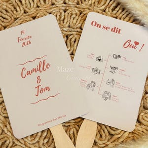 Peut inclure: Deux éventails de mariage rose clair avec des manches en bois. L'un porte la date "14 Février 2026" et les noms "Camille & Tom" en script rouge. L'autre affiche un programme de mariage avec des illustrations et la phrase "On se dit Oui!" en rouge.