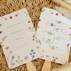 Peut inclure: Deux éventails de mariage avec des motifs floraux et des manches en bois. Les éventails présentent les noms "Mathilde et Paul" et la date "20.08.2026". Un éventail affiche un programme de mariage avec des illustrations.