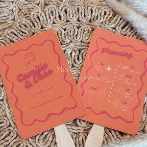 May include: Two orange wedding fans with pink text and wavy borders. One fan reads "Camille & Theo" and the date "12 Avril 2025". The other fan has a wedding timeline. Both fans have wooden handles.