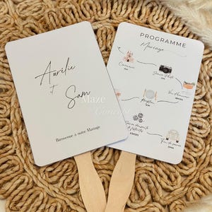 Peut inclure: Deux éventails de mariage blancs avec des manches en bois. L'un porte les noms "Aurélie & Sam" et "Bienvenue à notre Mariage". L'autre affiche un programme de mariage avec des illustrations et le texte "Programme Mariage".