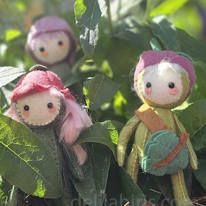 May include: Three handmade felt dolls nestled among green leaves and purple flowers. The dolls have round faces, button eyes, and colorful hats. One doll has a red hat and pink hair, another has a purple hat, and the third has a green outfit and a green satchel.