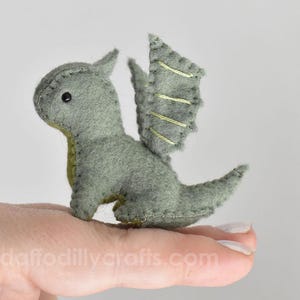 May include: A small, sage green felt dragon plush toy featuring visible dark grey stitching along the edges, a single black bead eye, and wings detailed with light yellow embroidered lines. The creature has a pointed tail and small horns.
