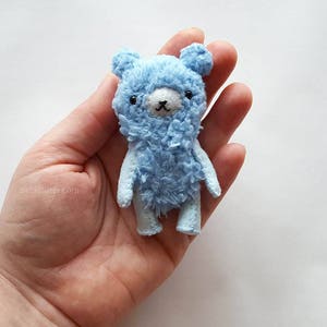 May include: A small, light blue teddy bear with black eyes and a white nose. The bear is made of soft, fluffy fabric and has a slightly distressed look.