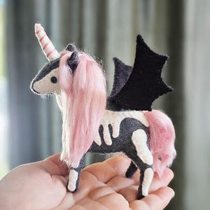 Puede incluir: Un unicornio de fieltro hecho a mano con pelo y cola rosa, un cuerno a rayas y alas de murciélago negras. El unicornio tiene un cuerpo gris con marcas blancas en forma de hueso. Un artículo decorativo único y caprichoso.