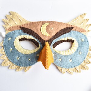 Op de afbeelding: Een vilt uilenmasker in bruin, beige en blauw. Het masker heeft een halve maan en sterren geborduurd op het voorhoofd. Het masker heeft een snavel en ooggaten.
