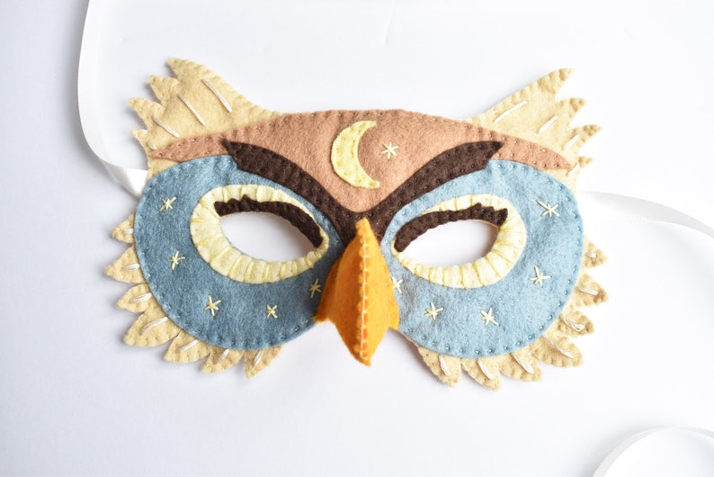 Op de afbeelding: Een vilt uilenmasker in bruin, beige en blauw. Het masker heeft een halve maan en sterren geborduurd op het voorhoofd. Het masker heeft een snavel en ooggaten.