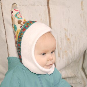 Op de afbeelding: Een witte fleece baby muts met een puntige, kleurrijke stoffen top. De muts heeft een flap die de oren en nek bedekt. De stoffen top heeft een bloemenpatroon en de tekst "www.delilahiris.com".