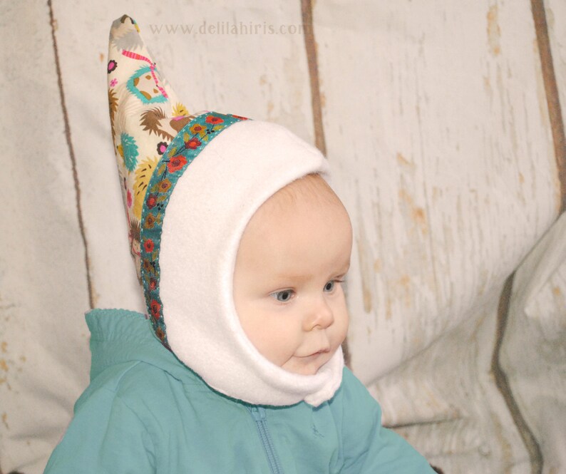 Op de afbeelding: Een witte fleece baby muts met een puntige, kleurrijke stoffen top. De muts heeft een flap die de oren en nek bedekt. De stoffen top heeft een bloemenpatroon en de tekst "www.delilahiris.com".