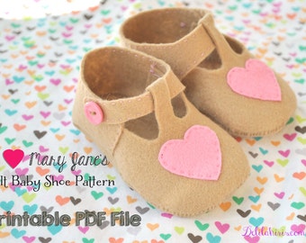 Op de afbeelding: Een paar bruine baby-schoentjes van vilt met roze hartjesaccenten. De schoentjes hebben een T-band ontwerp en zijn versierd met een knoop aan de voorkant. De tekst "I ♥ Mary Jane's Felt Baby Shoe Pattern Printable PDF File" is zichtbaar in de afbeelding.