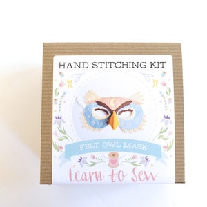 Op de afbeelding: Een bruine kartonnen doos met een wit label waarop "HAND STITCHING KIT" en "FELT OWL MASK" staat, met een kleurrijke illustratie van een uilenmasker. Het label heeft ook de tekst "Learn to Sew" in roze letters.