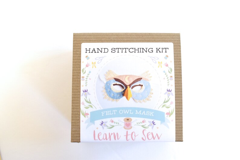Op de afbeelding: Een bruine kartonnen doos met een wit label waarop "HAND STITCHING KIT" en "FELT OWL MASK" staat, met een kleurrijke illustratie van een uilenmasker. Het label heeft ook de tekst "Learn to Sew" in roze letters.