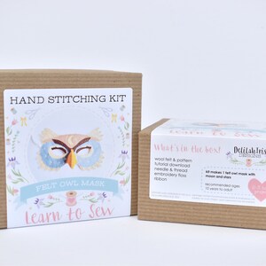 Op de afbeelding: Een bruine kartonnen doos met een kleurrijk label waarop staat "Hand Stitching Kit Felt Owl Mask Learn to Sew". Het label toont een illustratie van een blauw en bruin uilenmasker met een gele snavel. De doos staat naast een kleinere bruine kartonnen doos met een label waarop staat "What's in the box! Delilah Iris Designs". Het label somt de inhoud van de kit op, waaronder wolvilt, een patroon, een tutorial-download, een naald en draad, borduurgaren en lint. Het label zegt ook "Kit makes 1 felt owl mask with moon and stars".