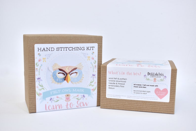 Op de afbeelding: Een bruine kartonnen doos met een kleurrijk label waarop staat "Hand Stitching Kit Felt Owl Mask Learn to Sew". Het label toont een illustratie van een blauw en bruin uilenmasker met een gele snavel. De doos staat naast een kleinere bruine kartonnen doos met een label waarop staat "What's in the box! Delilah Iris Designs". Het label somt de inhoud van de kit op, waaronder wolvilt, een patroon, een tutorial-download, een naald en draad, borduurgaren en lint. Het label zegt ook "Kit makes 1 felt owl mask with moon and stars".
