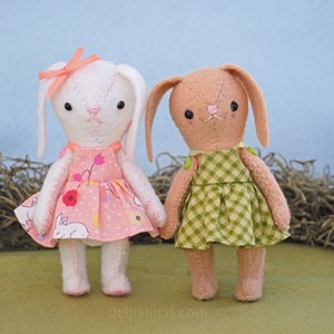 May include: Two handmade felt bunny dolls wearing dresses. The bunny on the left is white with a pink dress and a pink bow. The bunny on the right is brown with a green gingham dress.