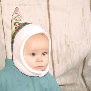 Op de afbeelding: Een baby draagt ​​een witte capuchon met een kleurrijke, gepatroneerde rand. De capuchon heeft een puntige top en de baby draagt ​​een tealblauwe jas.