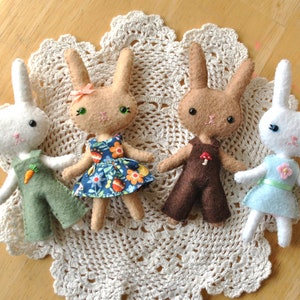 May include: Four handmade felt bunny dolls dressed in different outfits. The bunnies are sitting on a white crocheted doily. One bunny is wearing a green overall with a carrot, one is wearing a blue floral dress, one is wearing a brown overall with a mushroom, and one is wearing a light blue dress with a green ribbon.
