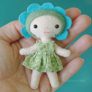 May include: A small felt doll with a cream-colored body, wearing a light green dress with a dark green floral pattern and a bright turquoise flower-shaped hat. The doll has black bead eyes and a simple stitched mouth. Text reads Delilahiris.com.