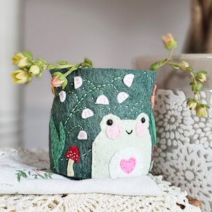May include: A green felt planter with a frog design. The frog is white with pink cheeks and a pink heart on its chest. The planter is decorated with green leaves and white flowers.