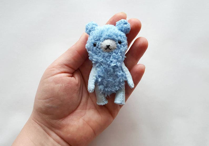 May include: A small, light blue teddy bear with black eyes and a white nose. The bear is made of soft, fluffy fabric and has a slightly distressed look.