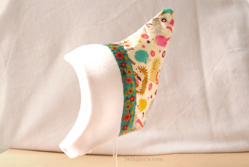 Op de afbeelding: Een witte baby muts met een puntige kroon en een kleurrijke stoffen rand met een egelpatroon. De rand heeft een turquoise, rode en gele kleurstelling.
