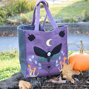May include: Purple felt tote bag with a black moth with a skull design, a crescent moon, and mushrooms. The bag is sitting on a tree stump with a small orange pumpkin.