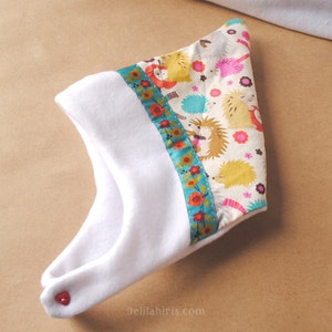 Op de afbeelding: Witte baby muts met een kleurrijke bies met een egelpatroon. De bies heeft een turquoise, rood en geel bloemenpatroon. De muts heeft een rode knoop aan de zijkant.