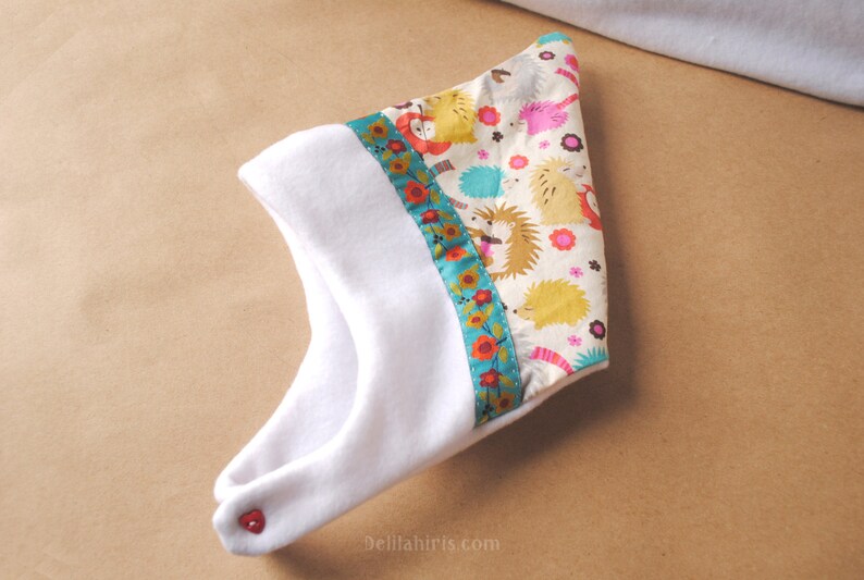 Op de afbeelding: Witte baby muts met een kleurrijke bies met een egelpatroon. De bies heeft een turquoise, rood en geel bloemenpatroon. De muts heeft een rode knoop aan de zijkant.