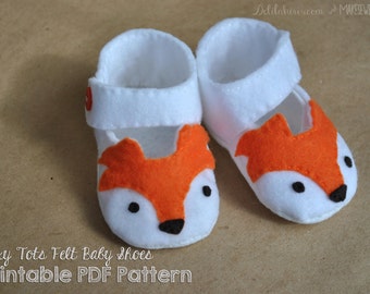 Op de afbeelding: Een paar witte baby-schoentjes van vilt met oranje vossengezichten. De schoentjes hebben een bandje over de bovenkant en zijn versierd met zwarte ogen en een zwarte neus.