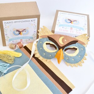 Op de afbeelding: Een handnaaikit voor het maken van een uilenmasker. De kit bevat viltstukken in bruin, beige, blauw en geel, borduurgaren en instructies. Het afgewerkte masker is een blauwe en bruine uil met een gele snavel en een halve maan op zijn voorhoofd.