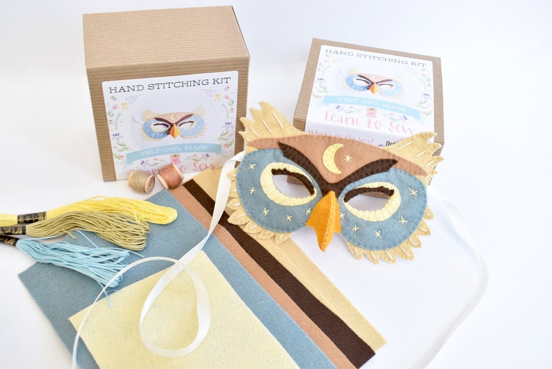 Op de afbeelding: Een handnaaikit voor het maken van een uilenmasker. De kit bevat viltstukken in bruin, beige, blauw en geel, borduurgaren en instructies. Het afgewerkte masker is een blauwe en bruine uil met een gele snavel en een halve maan op zijn voorhoofd.