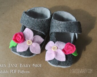 Op de afbeelding: Een paar grijze baby-schoentjes van vilt met roze en rode viltbloemen en groene viltbladeren. De schoentjes hebben een Mary Jane-bandje. De tekst "MARY JANE BABY SHOE Printable PDF Pattern" staat onderaan de afbeelding.