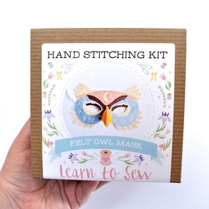Op de afbeelding: Een bruine kartonnen doos met een wit label waarop staat "HAND STITCHING KIT FELT OWL MASK Learn to Sew". Het label heeft een kleurrijke illustratie van een uilenmasker met een halve maan op zijn voorhoofd.