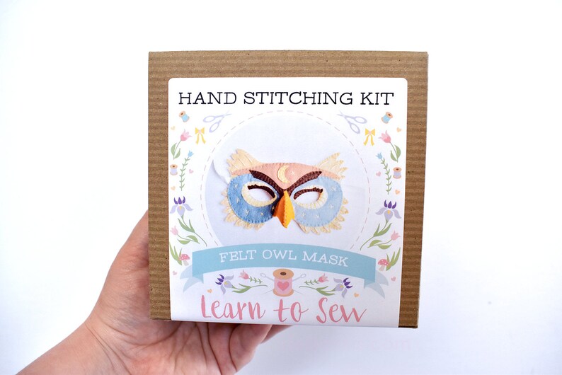 Op de afbeelding: Een bruine kartonnen doos met een wit label waarop staat "HAND STITCHING KIT FELT OWL MASK Learn to Sew". Het label heeft een kleurrijke illustratie van een uilenmasker met een halve maan op zijn voorhoofd.
