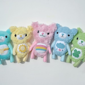 Peut inclure: Cinq ours en peluche de différentes couleurs, chacun avec un motif brodé différent. Les ours sont alignés de gauche à droite : bleu clair, jaune, rose, bleu clair et vert. Les motifs sont une étoile avec un arc-en-ciel, un soleil, un arc-en-ciel, un nuage avec des gouttes de pluie et un trèfle à quatre feuilles.